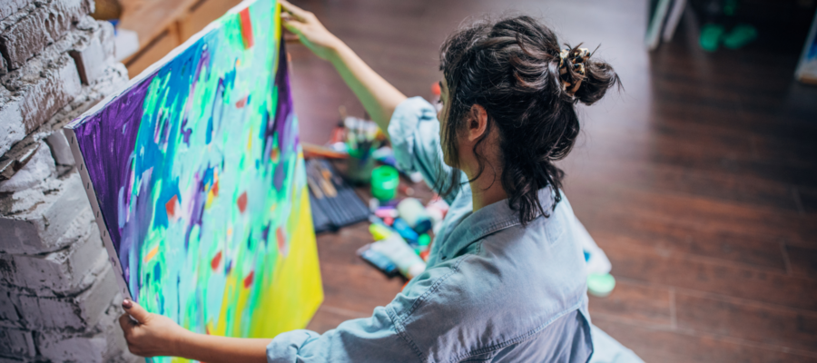 woman with brown hair in a bun holding up a colourful painting in her art studio
