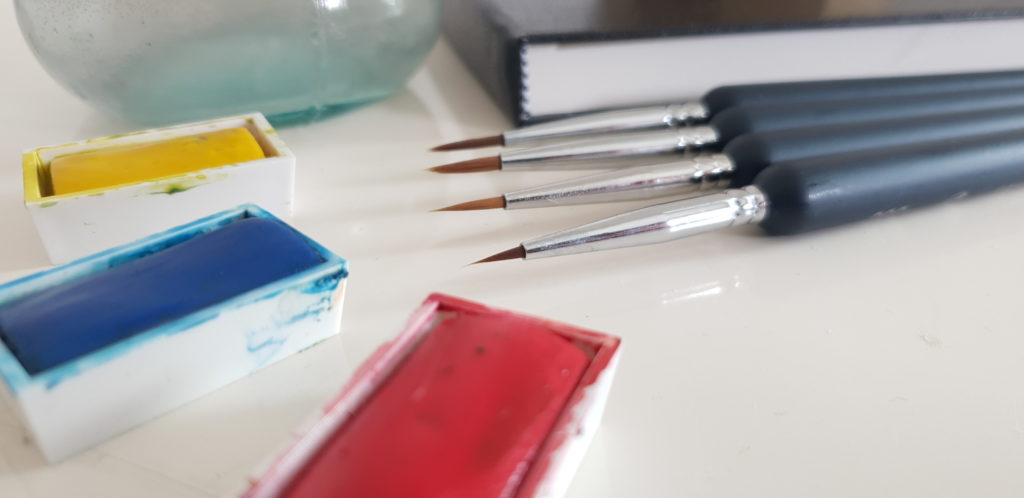 a set of four fine detail brushes lying next to primary watercolours yellow, blue and red