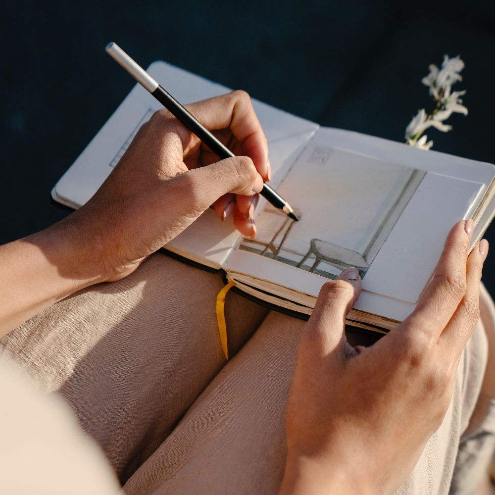 a woman using a graphite pencil to draw something in a notebook